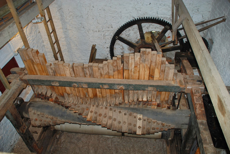 Old machinery of wooden blocks twisting round a metal cylinder with a tooth edged wheel in the background.