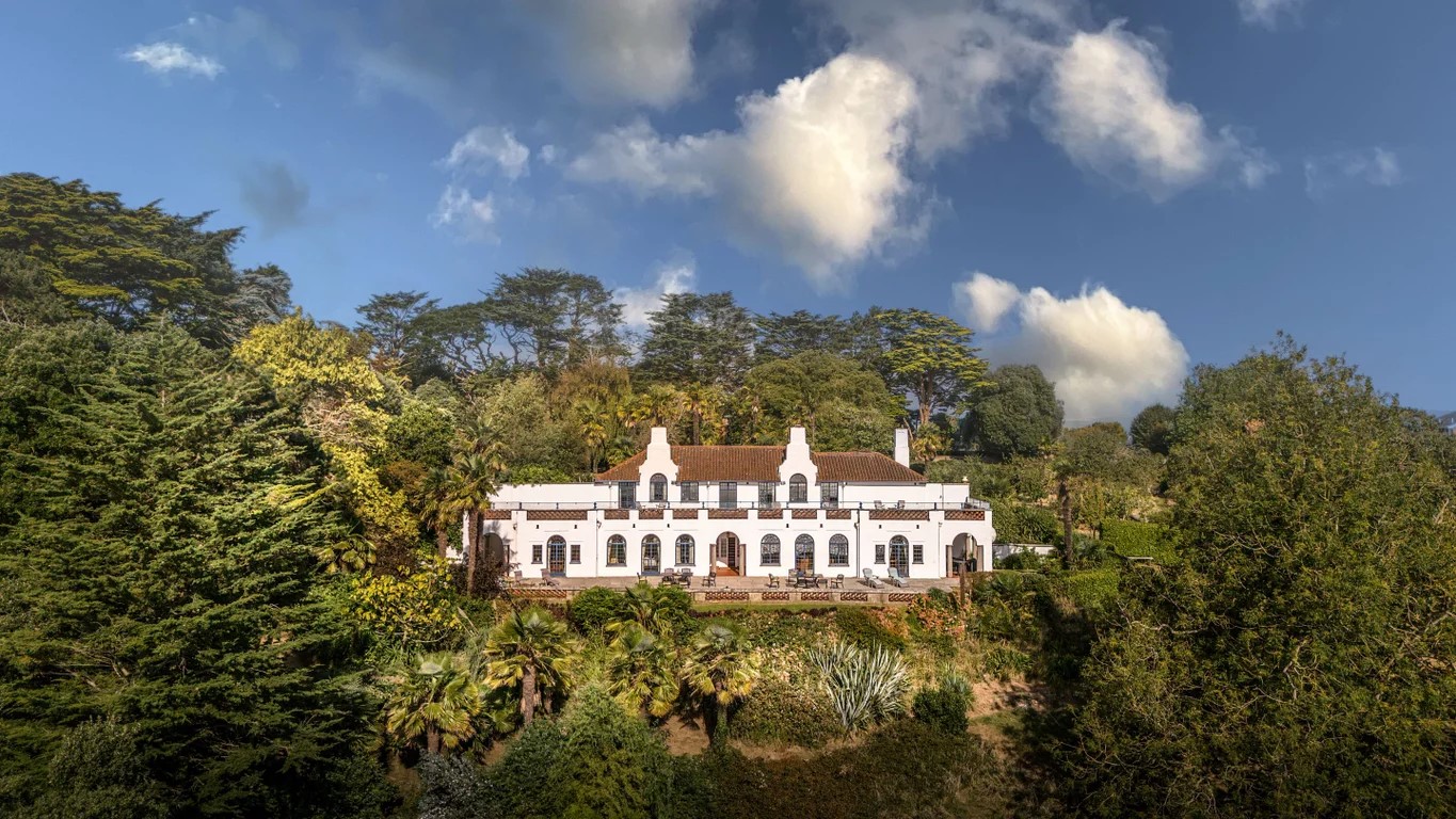 Long, two story white house set amid greenery on a hillside.