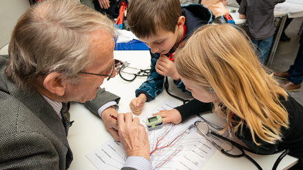 Older man and two children leaning across a desk with a medical diagram and pulse monitor.