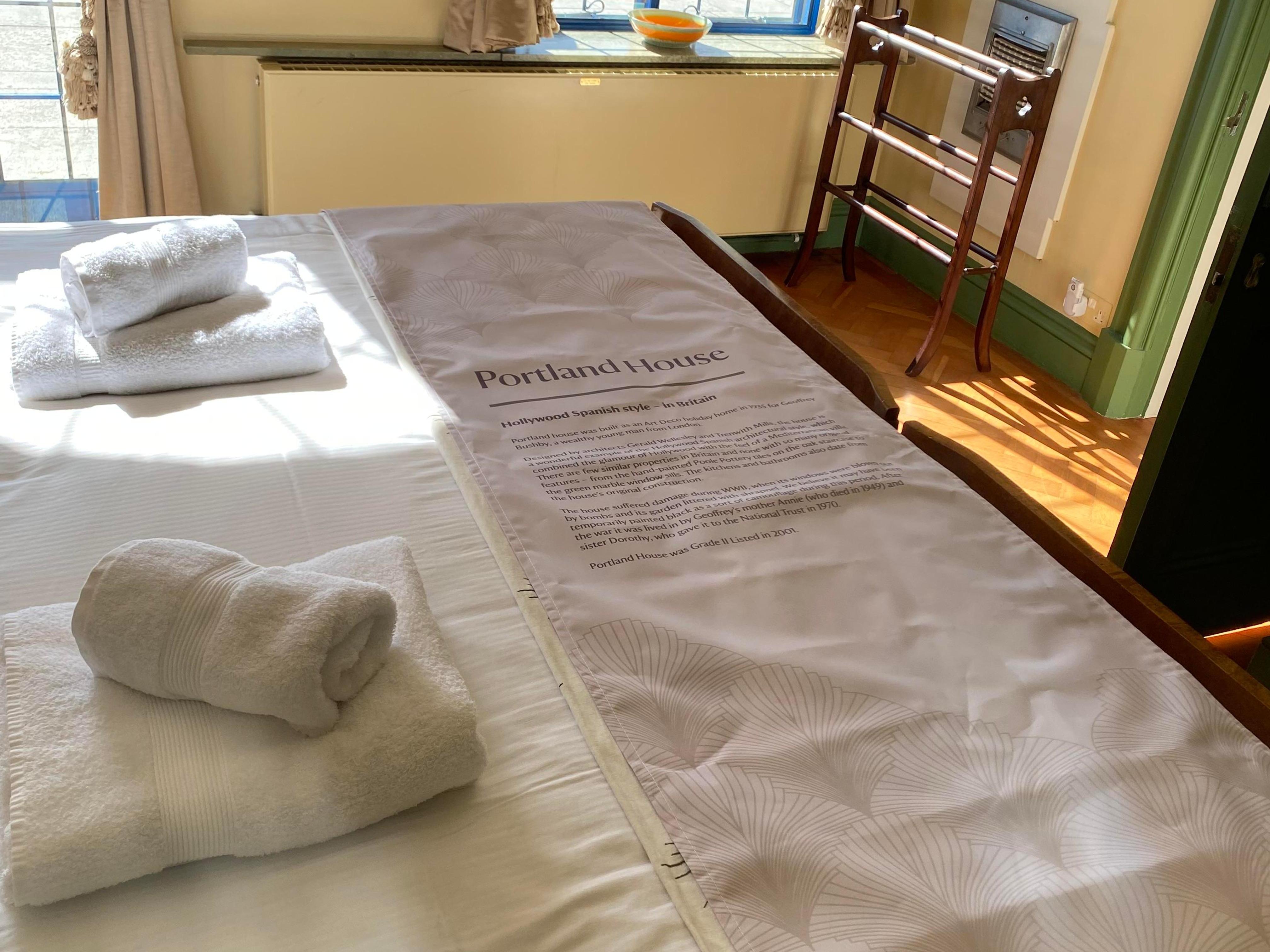 Folded towels and a long fabric sheet with text on it laid atop a double bed in a sunlit room.