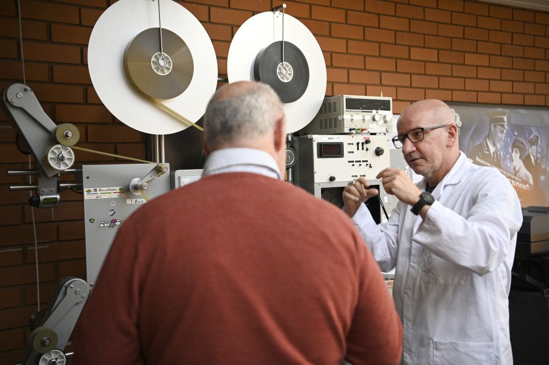 Two men, one in a white lab coat, standing by an old film reels.
