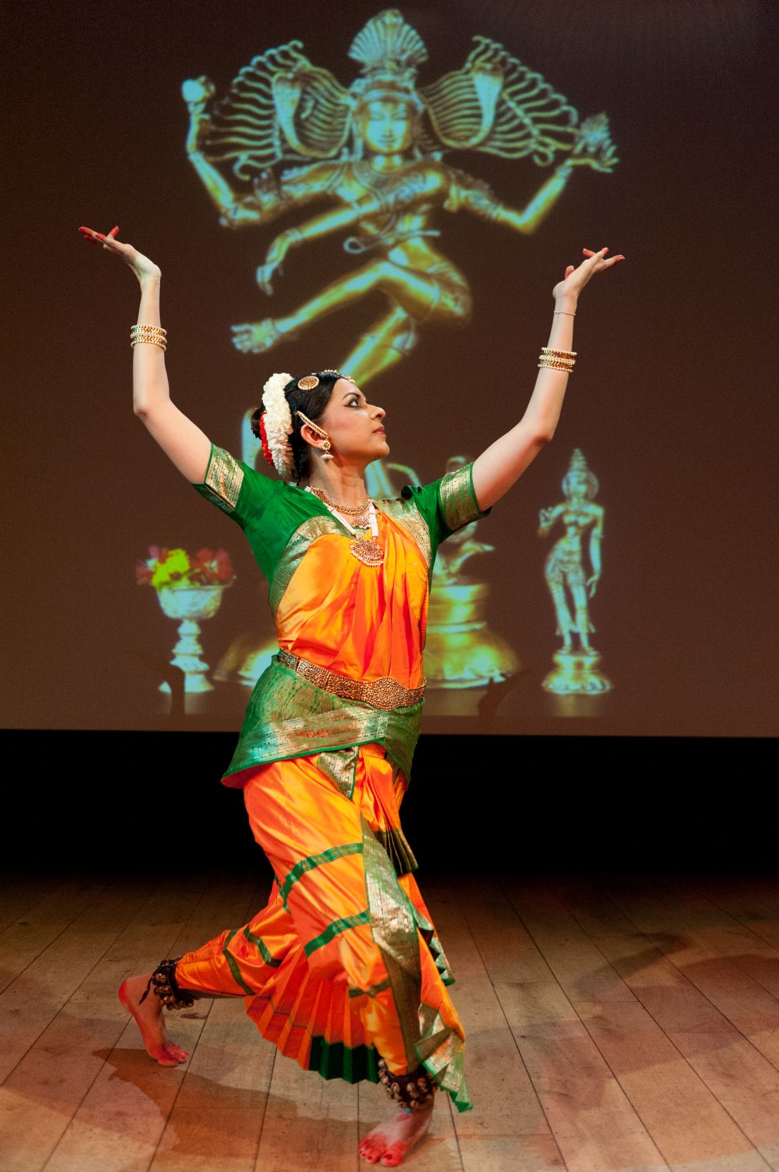 A woman in traditional orange and green Indian dress, flowers in her hair, dancing.
