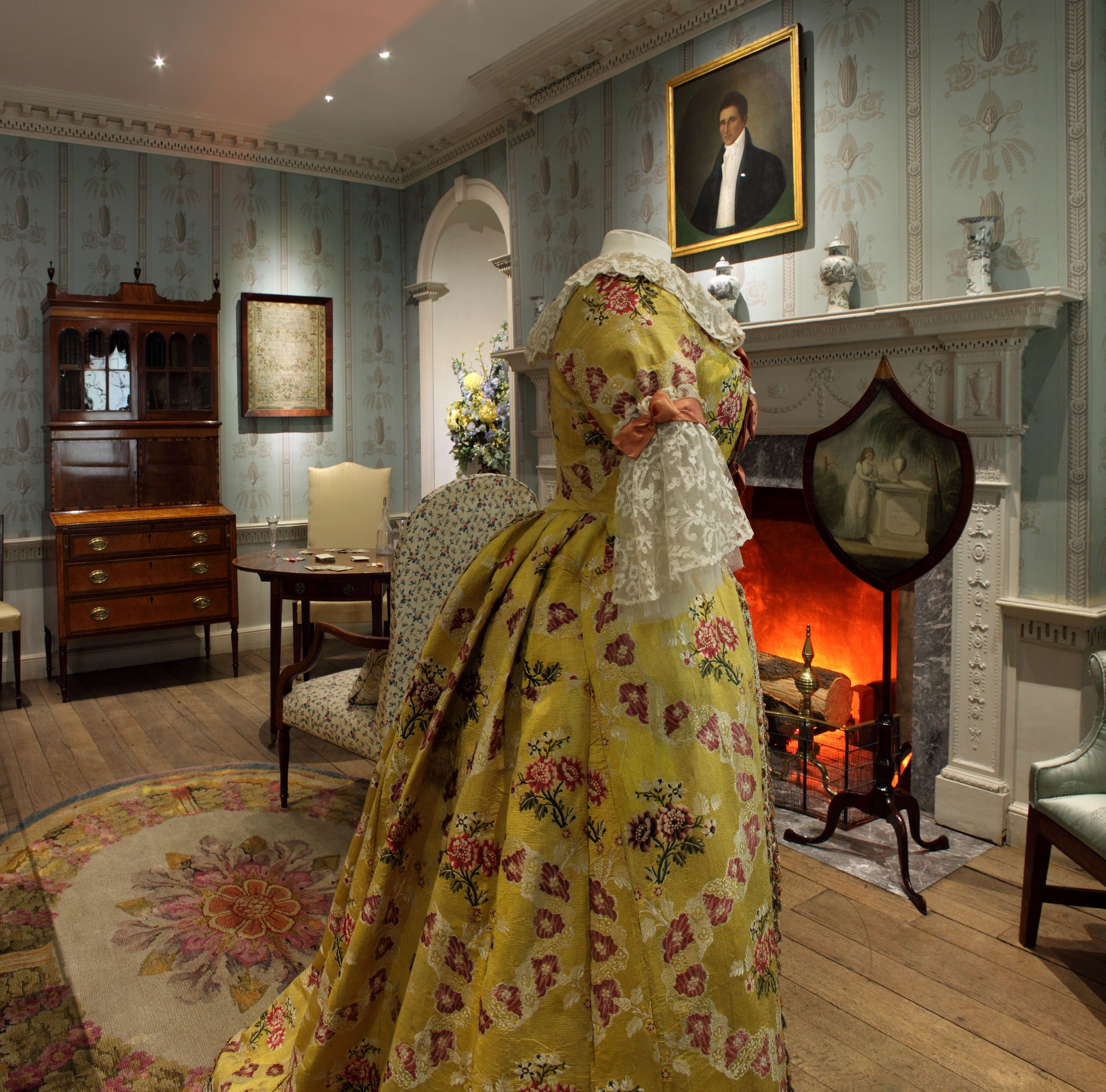 Fancy yellow dress on a costume stand in a historic room with blue patterned wallpaper and wooden floor.