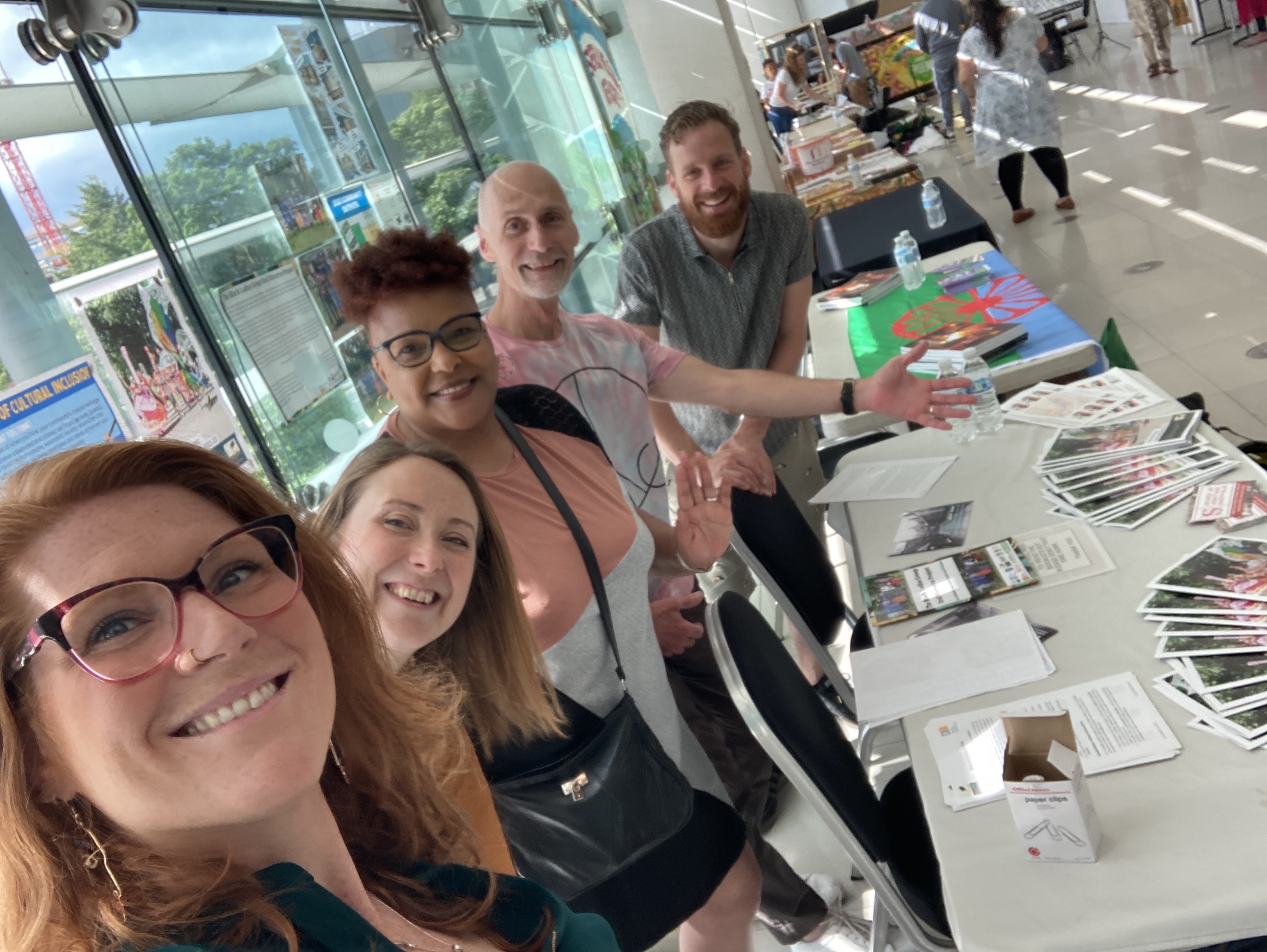 5 people stood in front of a glass wall and behind a table with papers on it in a large, sunny room.