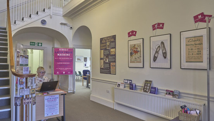 A women sat at a desk in the downstairs area of a building. To her left is the start of the staircase, to the right displays and HODs flags!