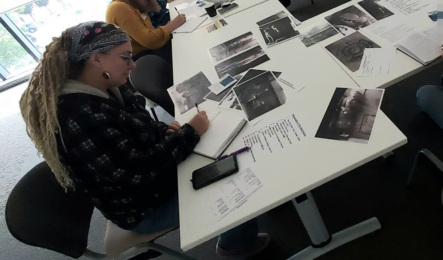 Woman with dreadlocks writing in a notebook sat at a table with assorted photo print outs scattered on it.