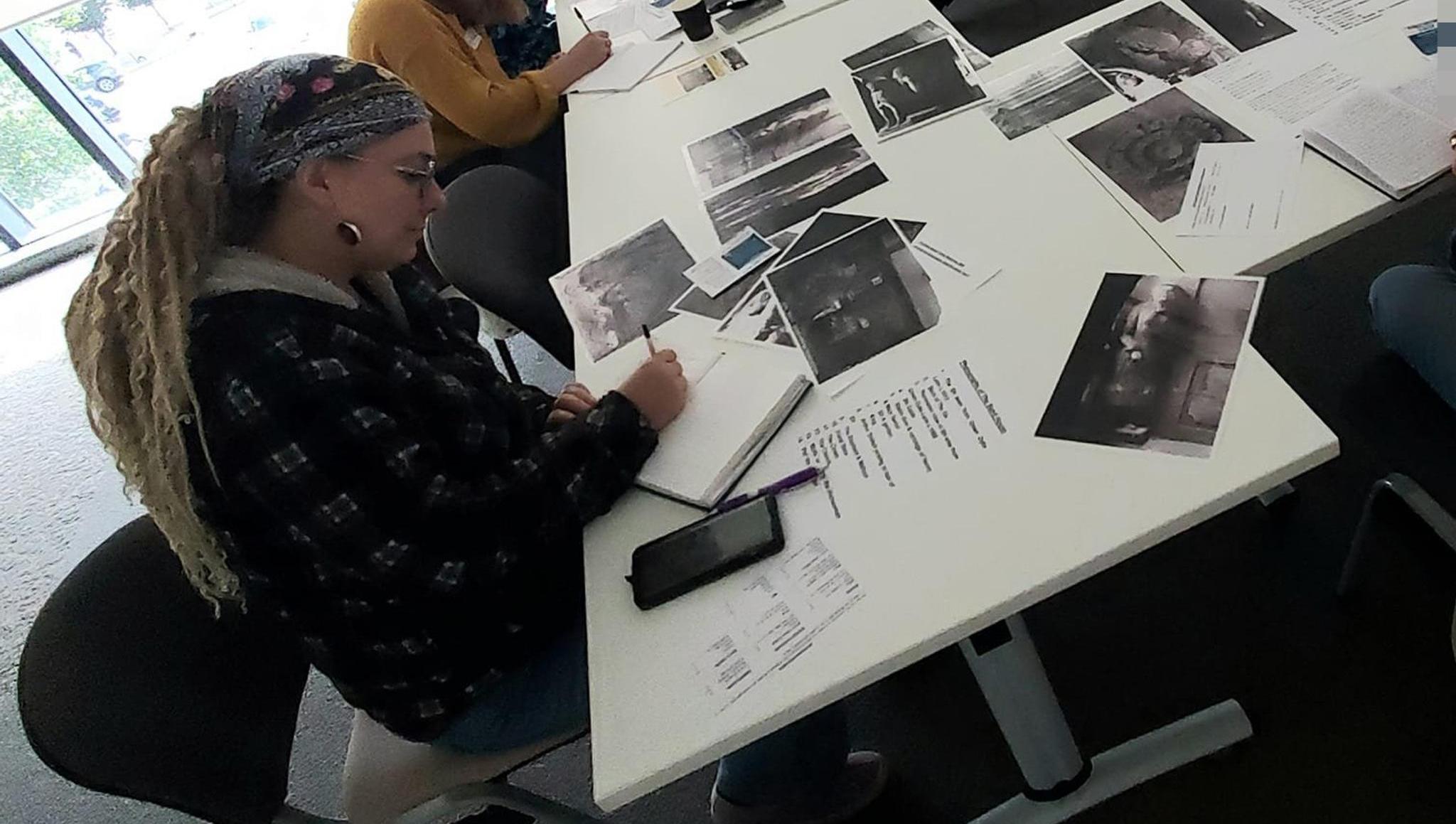 Woman with dreadlocks writing in a notebook sat at a table with assorted photo print outs scattered on it.