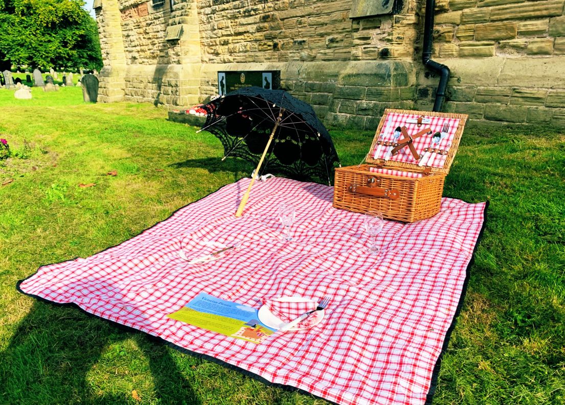 A red gingham picnic cloth sat on the grass at a grave yard. On top of the cloth is a wicker picnic basket, umbrella, a paper place and leaflet.