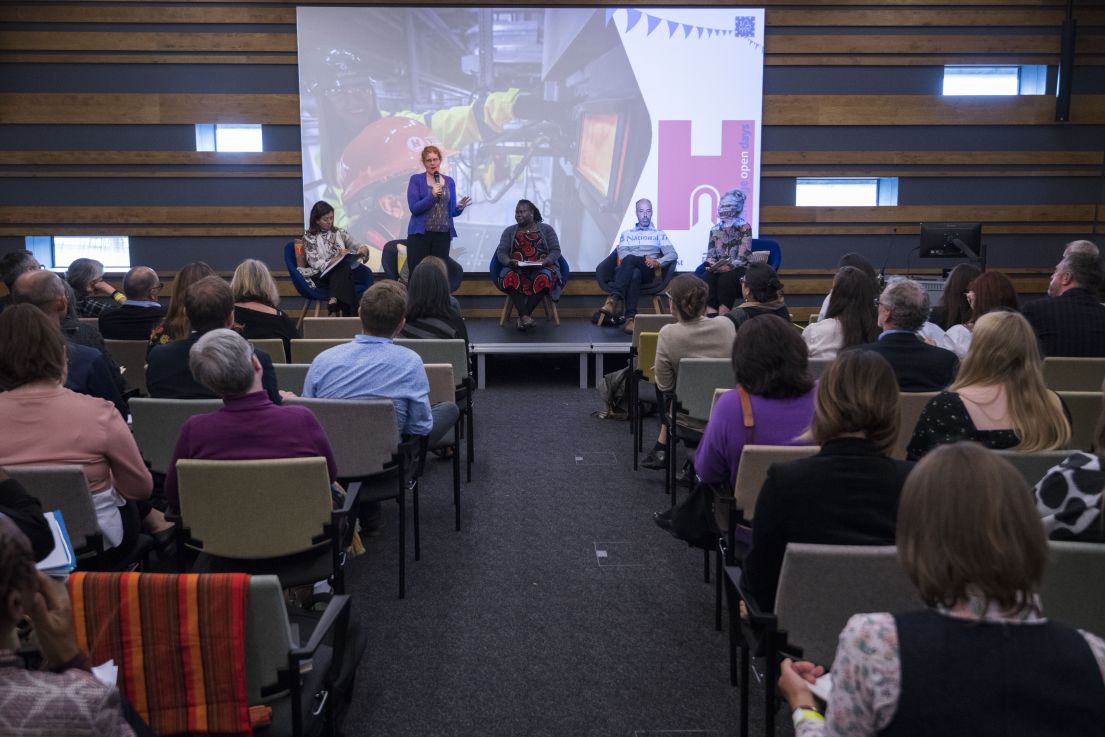 A group of people sat in rows of chairs listening to a panel present a presentation. On the stage a women with ginger hair is standing holding a mic.