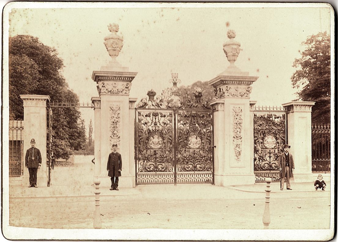 A decretive double metal gate with two white stone carved pillars either side. Then two single gates either side of that. 2 policemen standing guard.