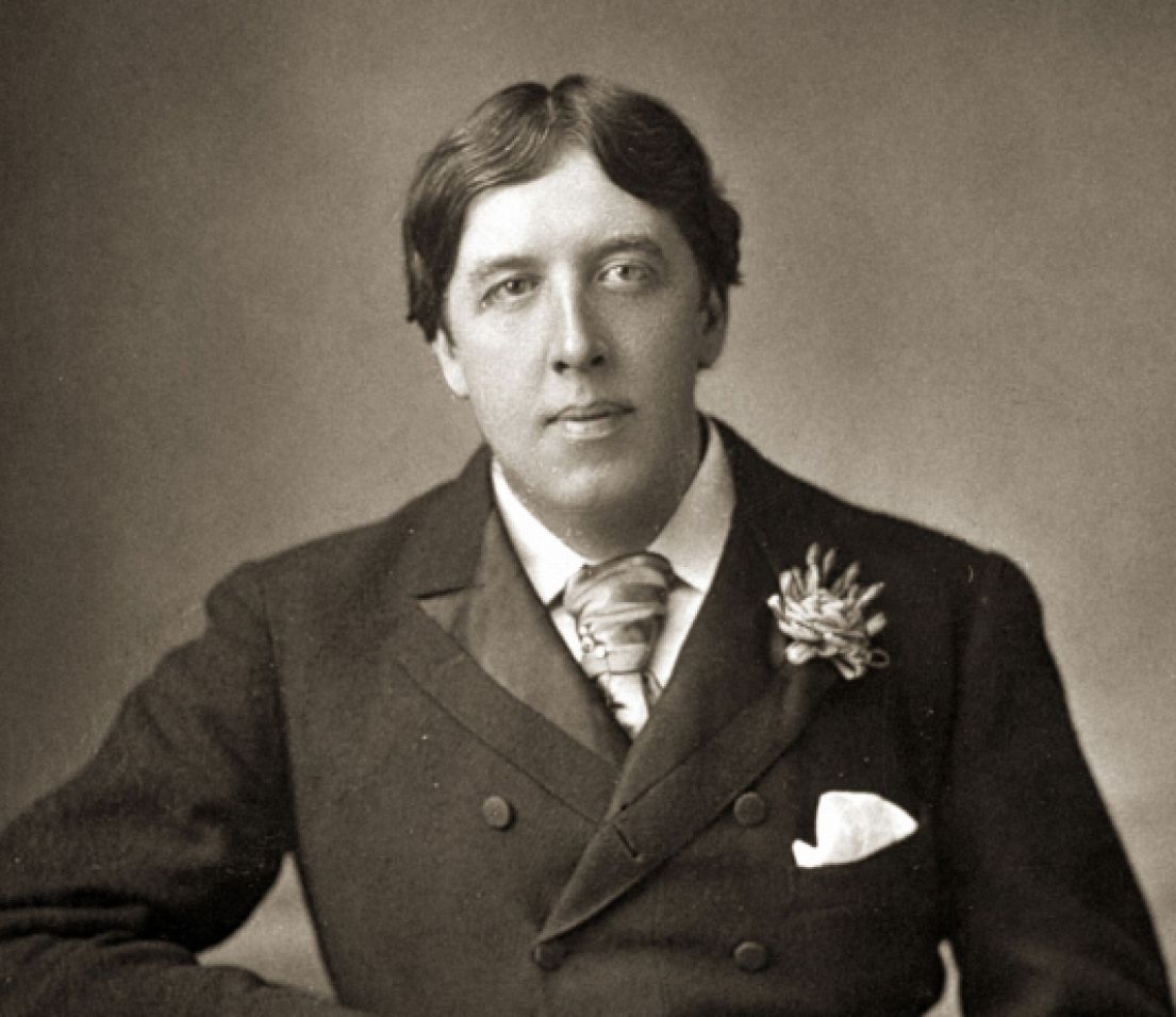 A black and white photo of a man. He has short hair with a side parting, wearing a smart suite, tie, pocket square as well as a flower in his lapel.