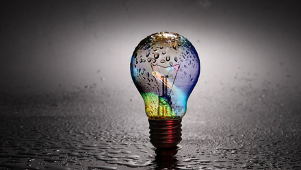 Lightbulb standing upright on wet grey surface. Reflecting rainbow colours from the water.