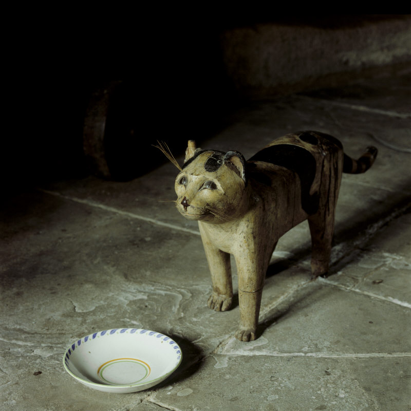 Wooden cat stands by china saucer on stone flagged floor.