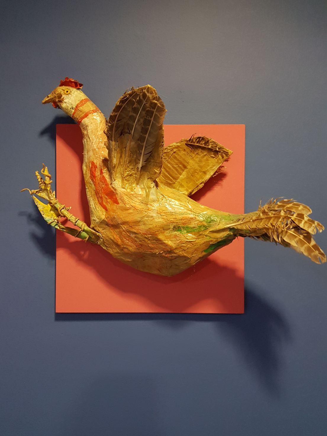 A colourful orange and yellow paper Mache cockerel attached to a grey wall. 