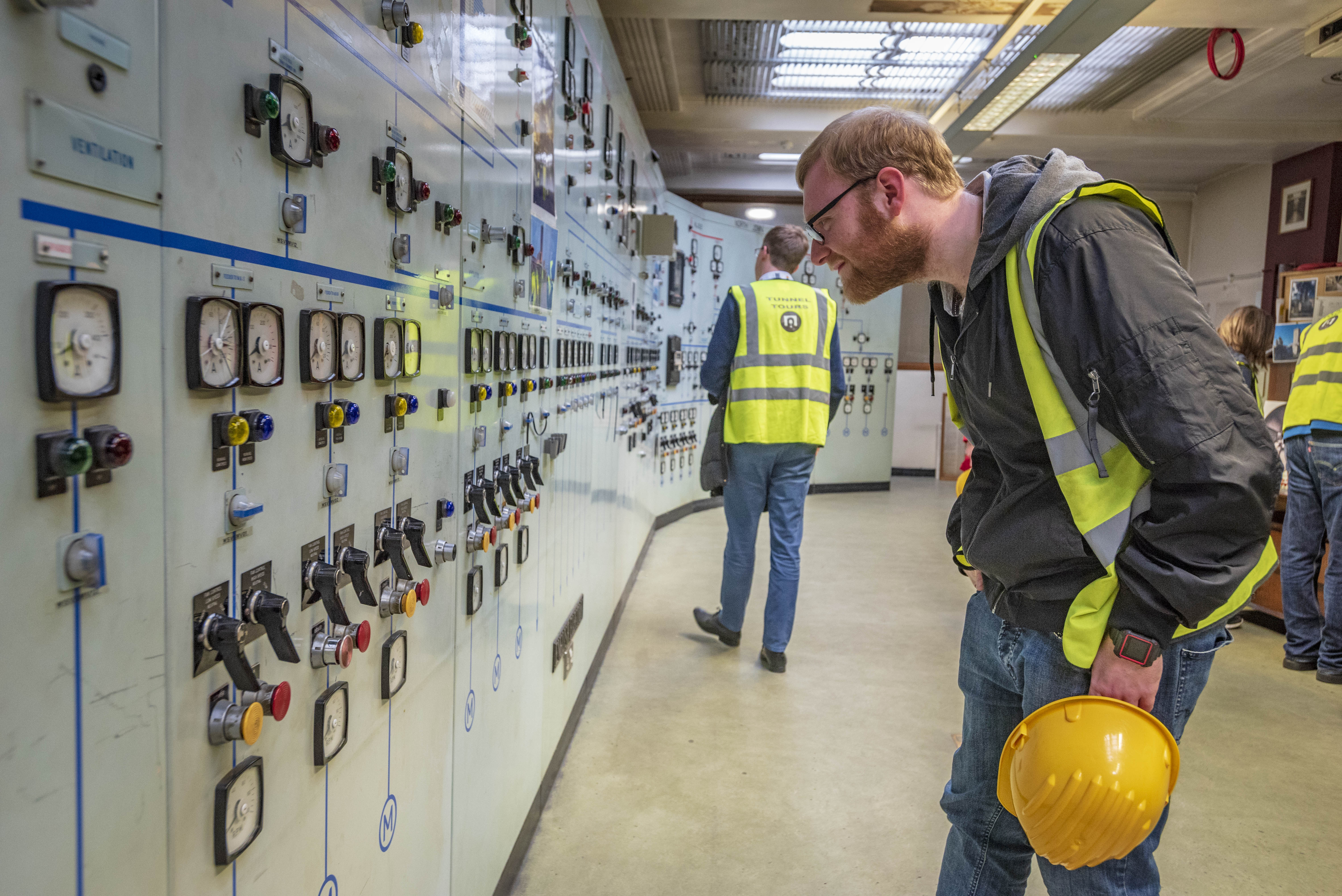 People in high vis tabards looking at a bank of grey machinery along a curving wall.