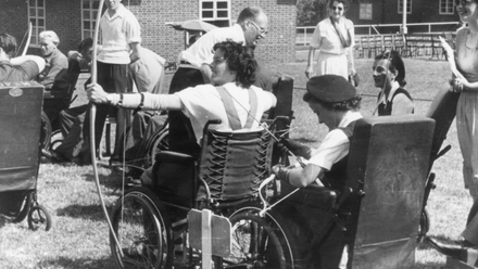 Old photograph of two women archers in wheelchairs on a grass field with a line of people watching behind them.