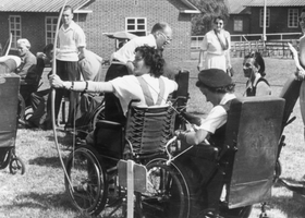 Old photograph of two women archers in wheelchairs on a grass field with a line of people watching behind them.