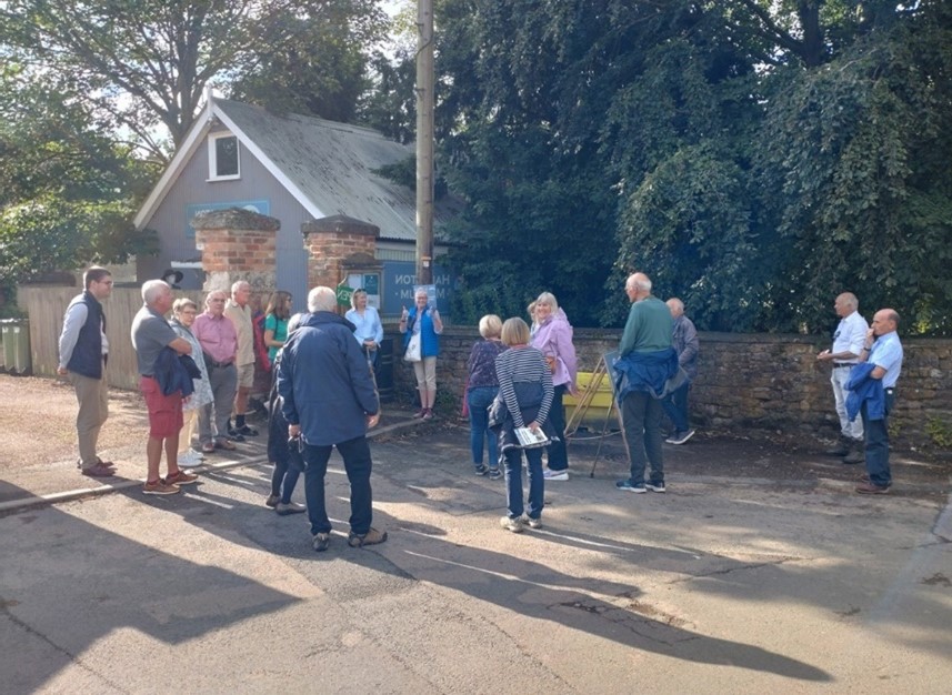 Group of people on a guided tour stopped on paved area beside a small building.