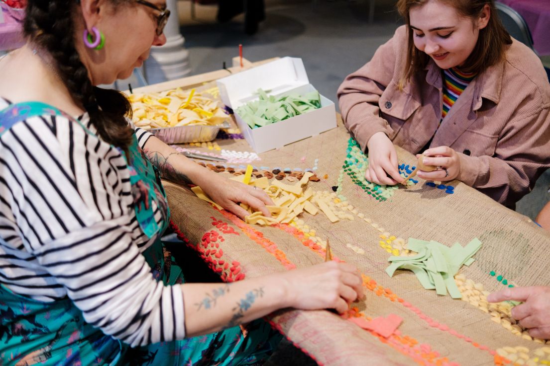 Two women sat at a table crafting - pulling coloured fabric through a hessian cloth.