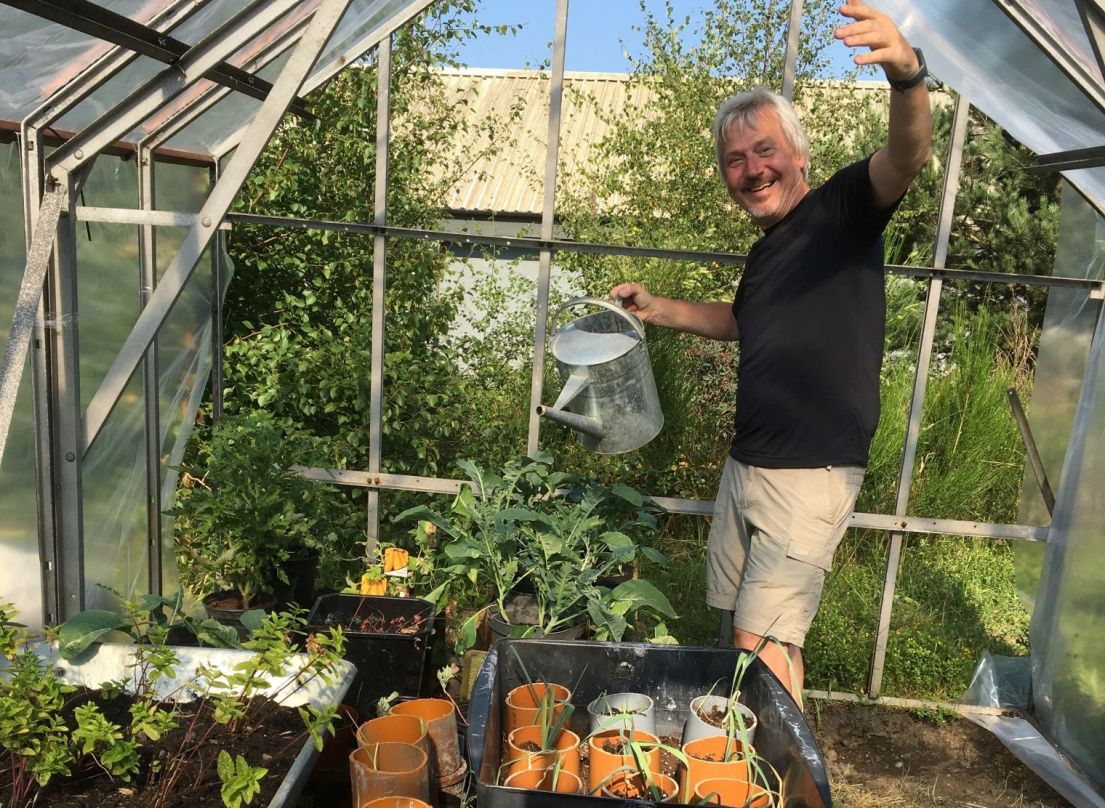 A man in a black t-shirt and cream cargo shorts holding a watering can, posing over his sprouting seedlings planted in pots or a wheelbarrow!