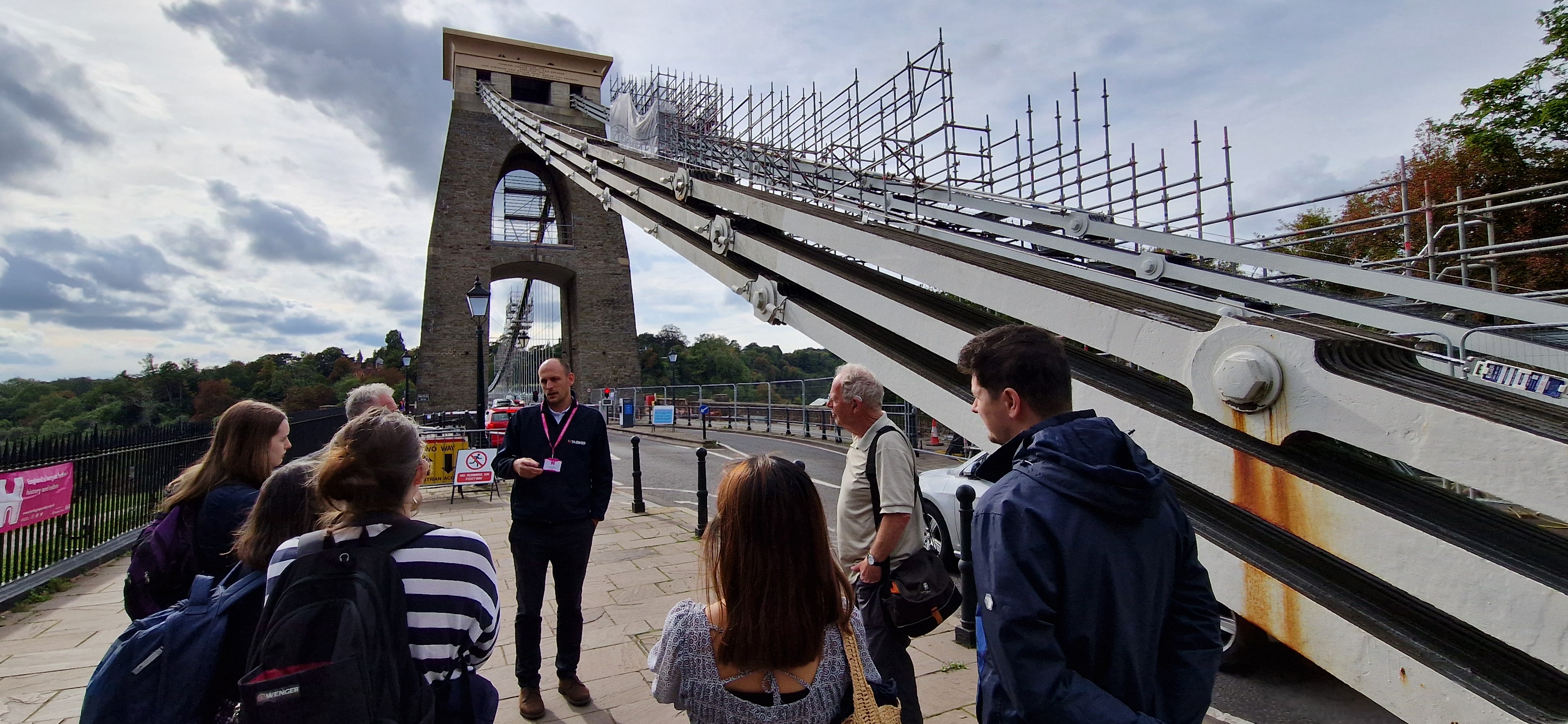 Man talking to a group of visitors stood on a pedestrianised bit of a large road bridge, with a suspension cable and scaffolding running up behind.