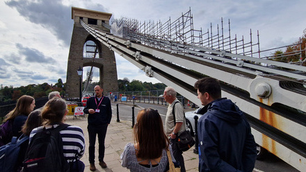 Man talking to a group of visitors stood on a pedestrianised bit of a large road bridge, with a suspension cable and scaffolding running up behind.