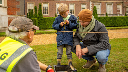 Older man in high vis showing a younger man and child a small object. Knelt on ground by a pit in front of a grand house.