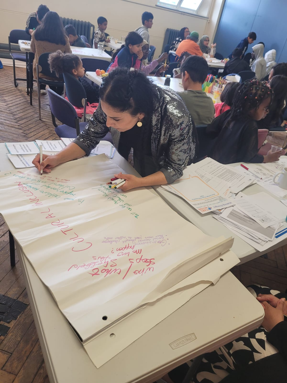 Woman leaning over flipchart paper on a table with several tables of children behind her.