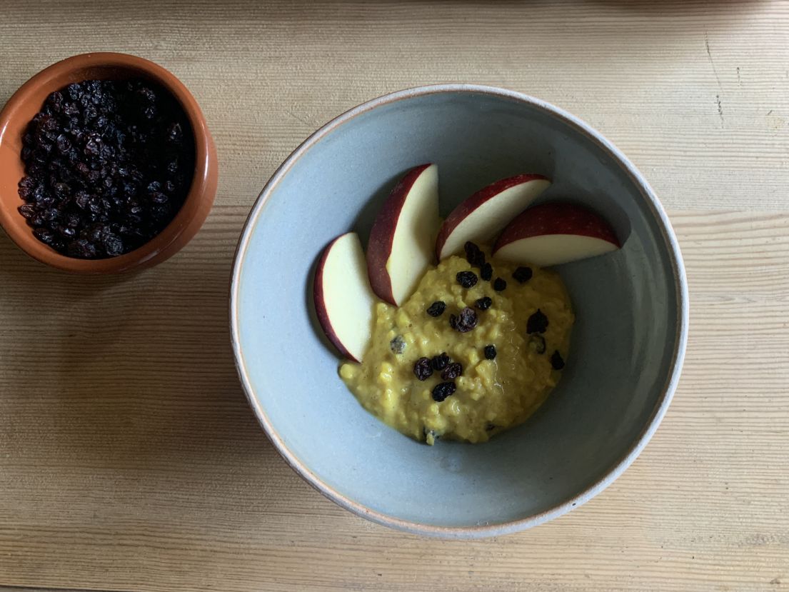 A blue bowl with yellow textured grained pudding. On the side are apples and the other currents.