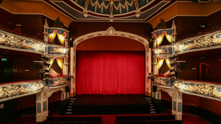 Ornate theatre auditorium with 2 tiers of gilded galleries and a red velvet curtain across the stage.