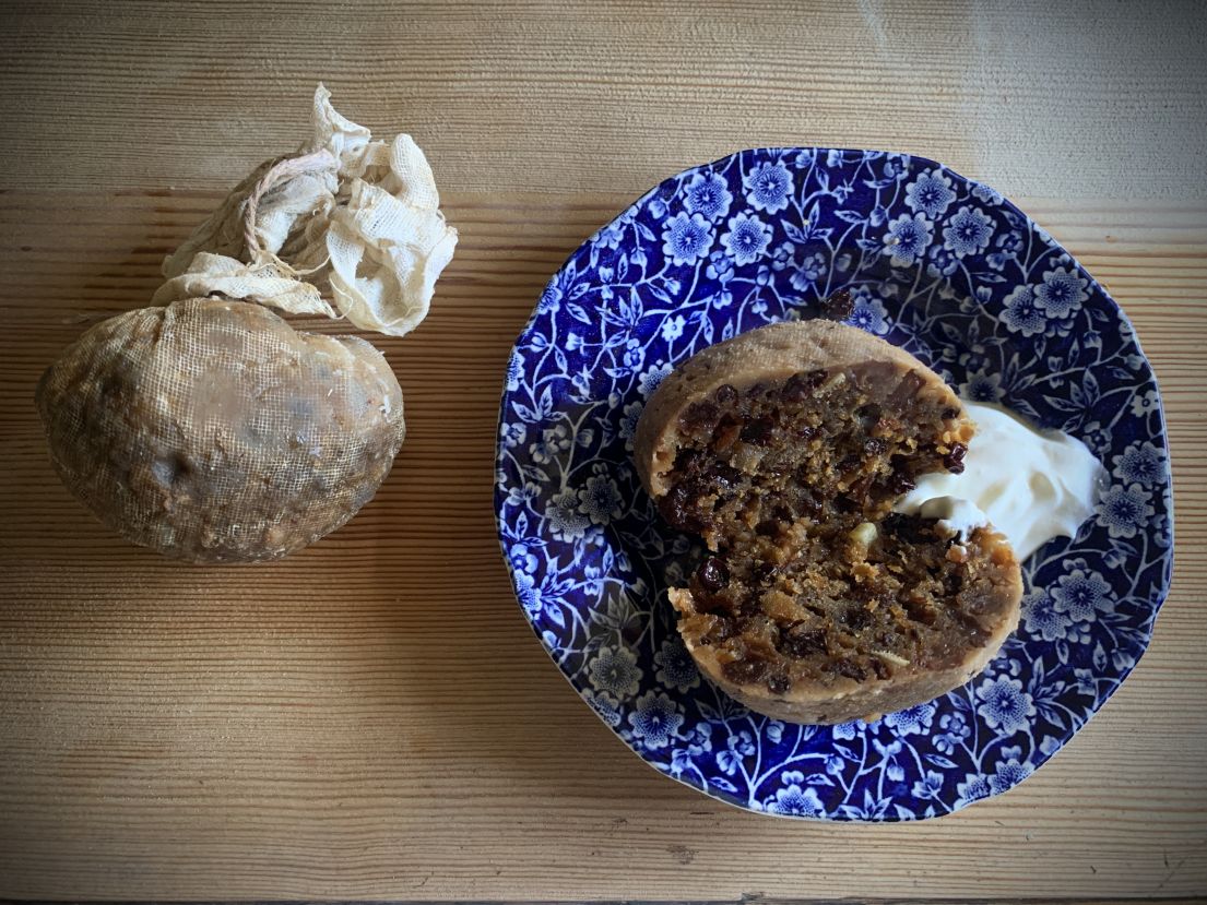 A figgy pudding. On the left shows the pudding mixture in a muslin cloth, the right is the pudding in a flower bowl with cream.