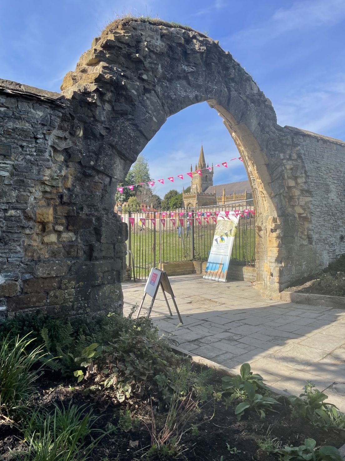 A medieval stone arch, which through can be seen a Abbey, with gardens adorned with pink 'HODs' bunting.