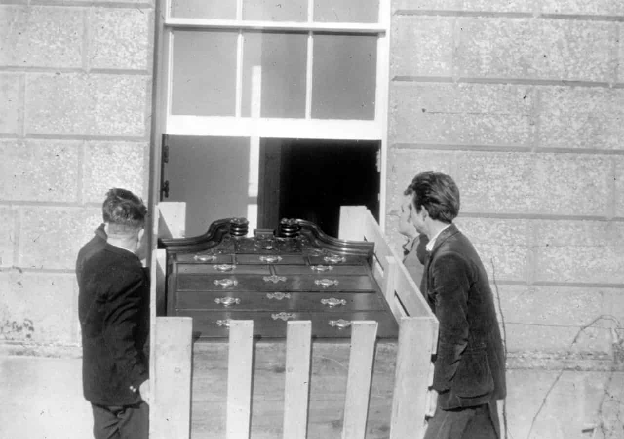 Photograph of four men lifting a chest of drawers through a window.