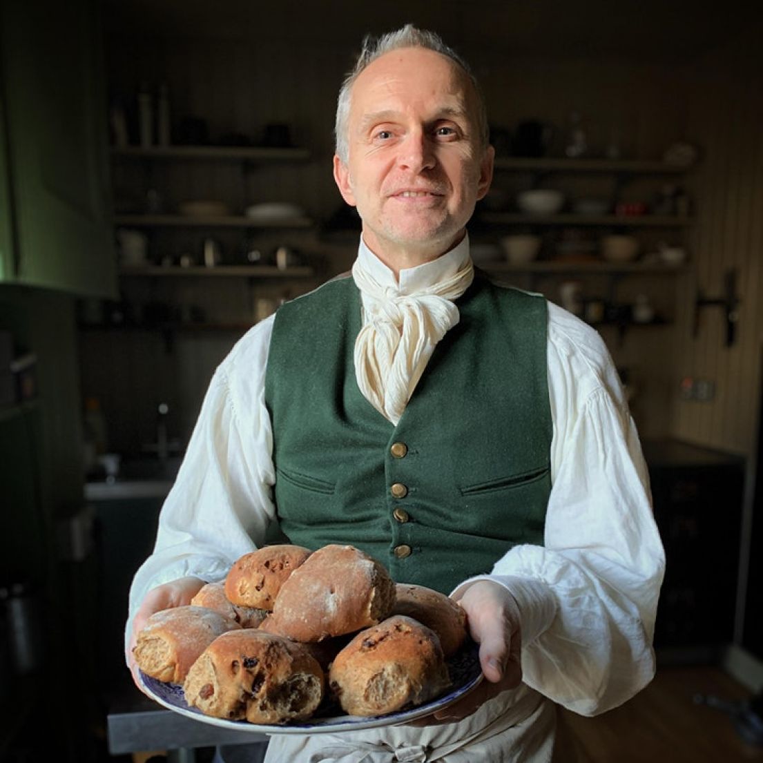 A man dressed in a regency shirt and dark green waistcoat holding a plate of English fruit buns.