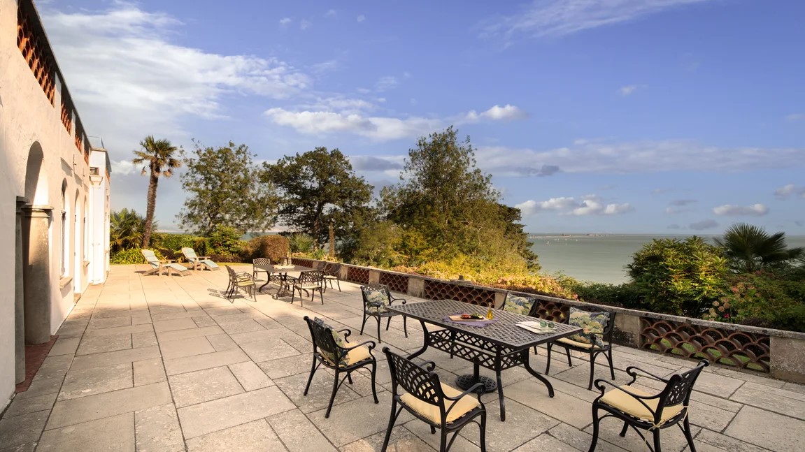 Chairs and tables on a stone flagged terrace with a view out to sea.