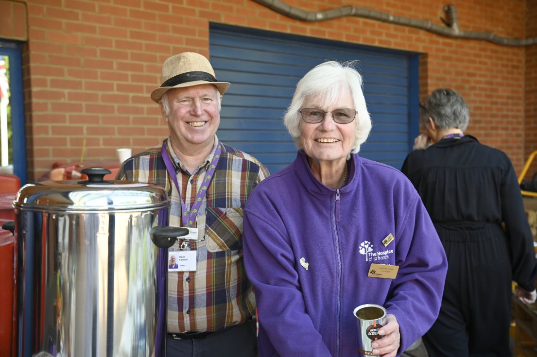 Two volunteers smiling, standing by a large flask of coffee to hand out to visitors.