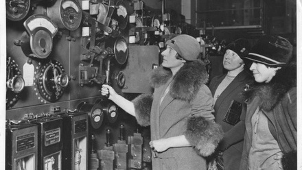 A black and white image of three women dressed in 1930's coats with fur trim and deep hats, examining dials in a factory.