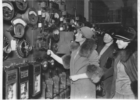 A black and white image of three women dressed in 1930's coats with fur trim and deep hats, examining dials in a factory.