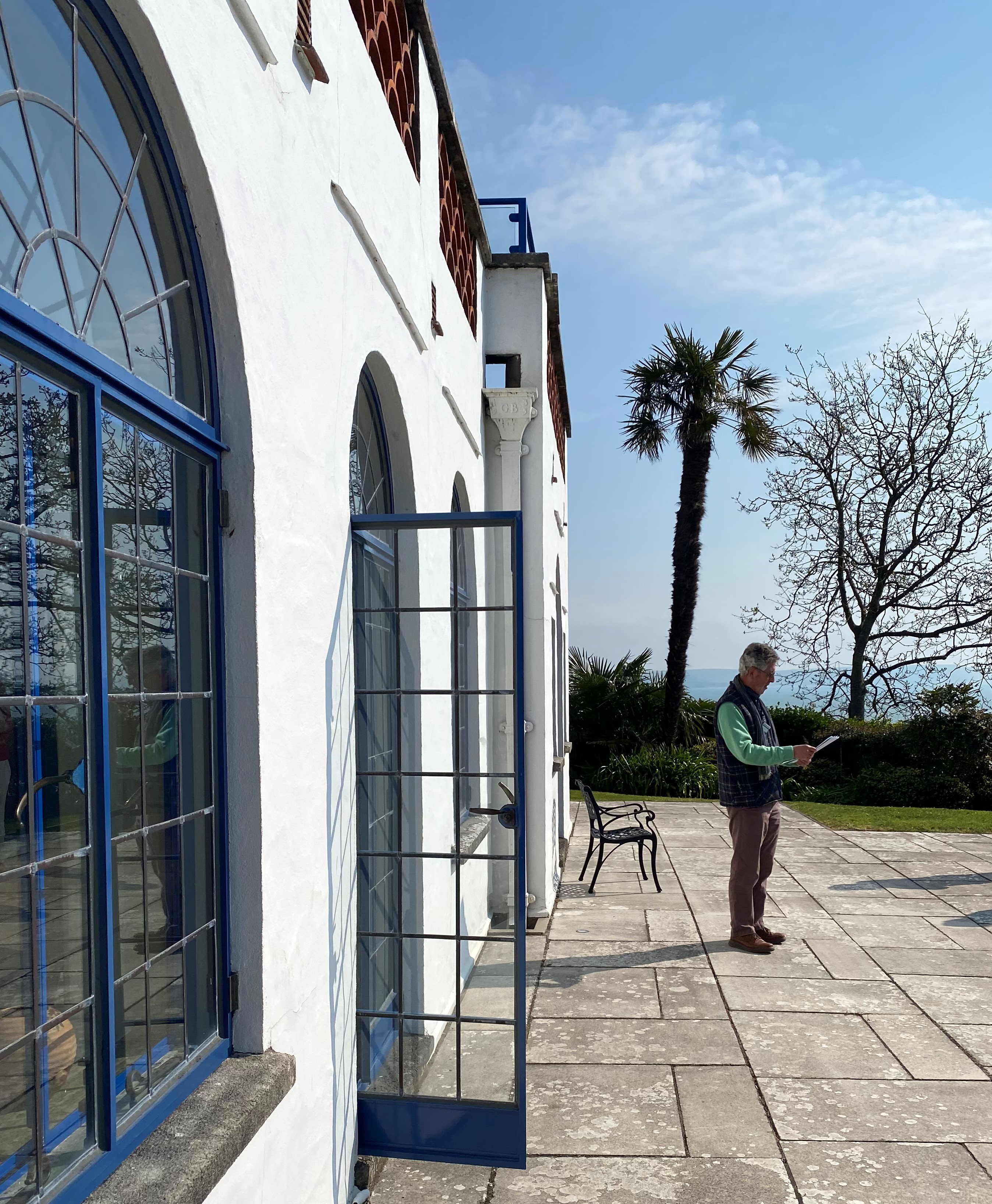 Man stood outside a white building with arched, blue framed windows, on a stone flagged terrace.