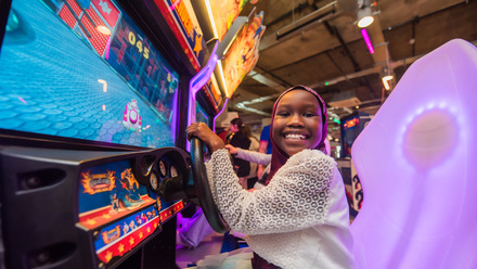 A photograph of a child smiling at the camera while playing a video game