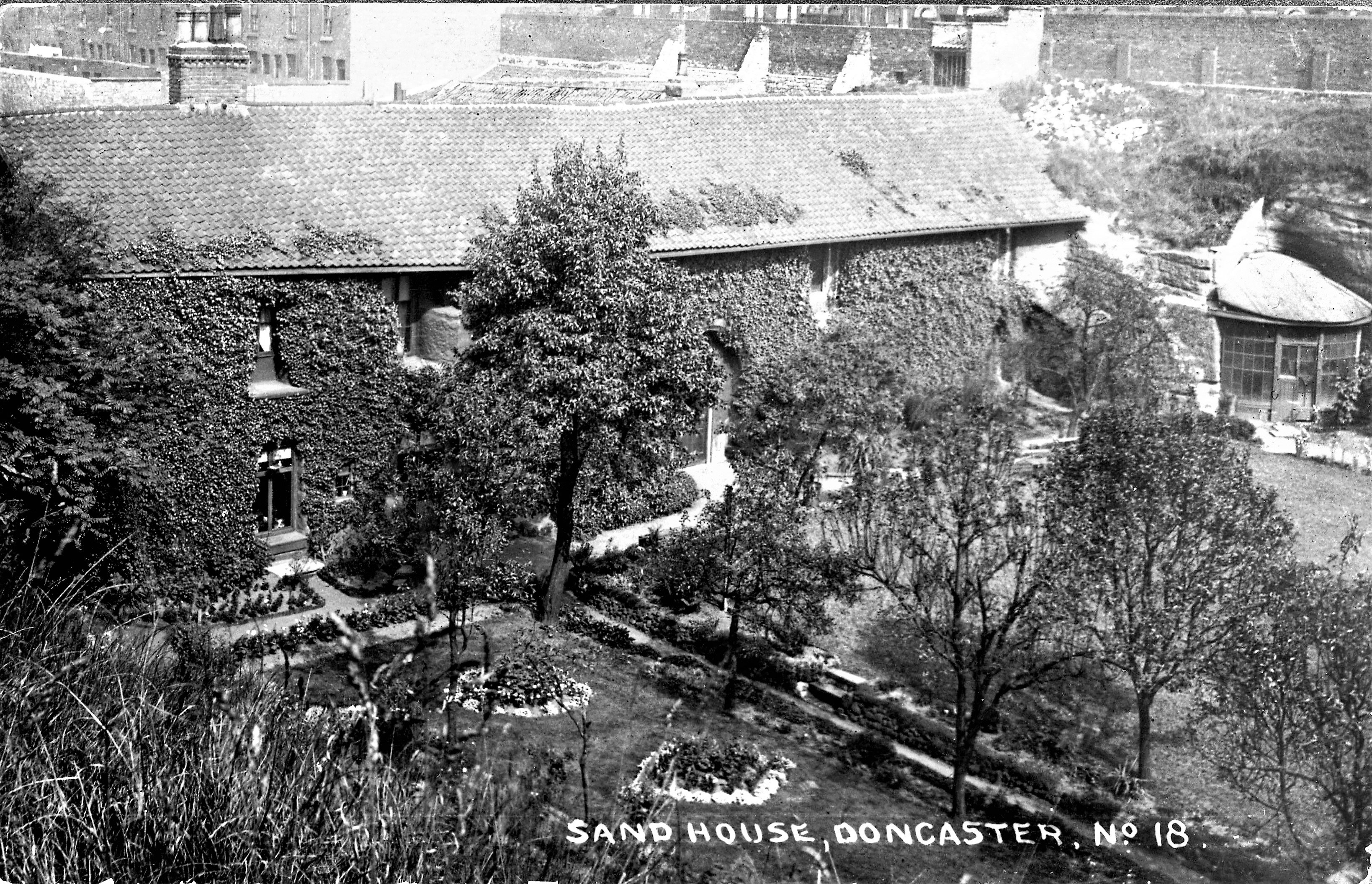 Old photograph of a long two story stone house covered in ivy with a garden in front.