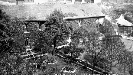 Old photograph of a long two story stone house covered in ivy with a garden in front.