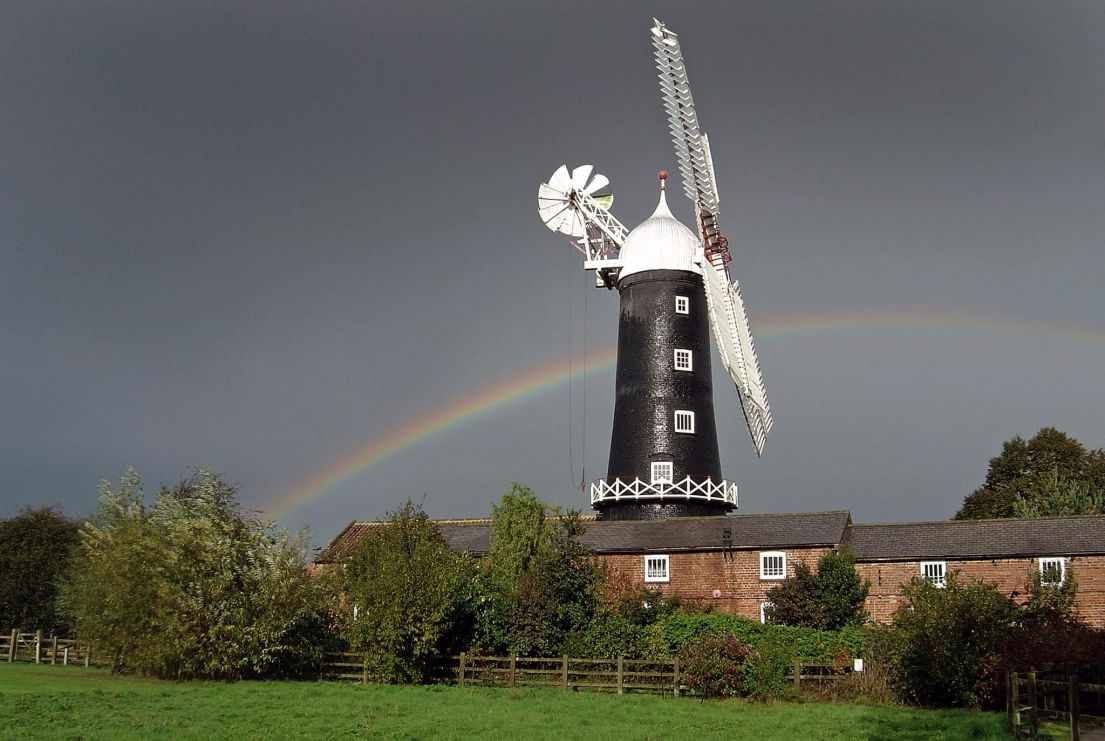 A black windmill with bright white sales. It is set in front of a dark overcast sky with a bright rainbow.