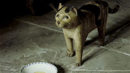 Wooden cat stands by china saucer on stone flagged floor.