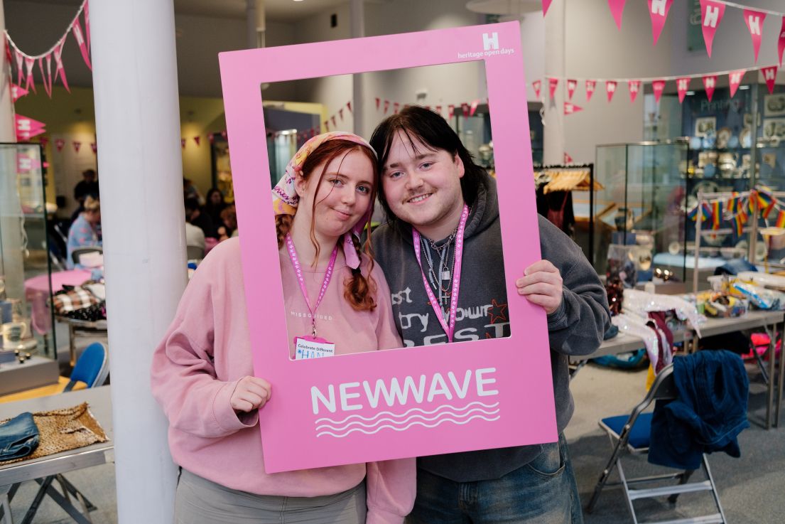Two young adults looking through a pink selfie frame in a room decorated with bunting.