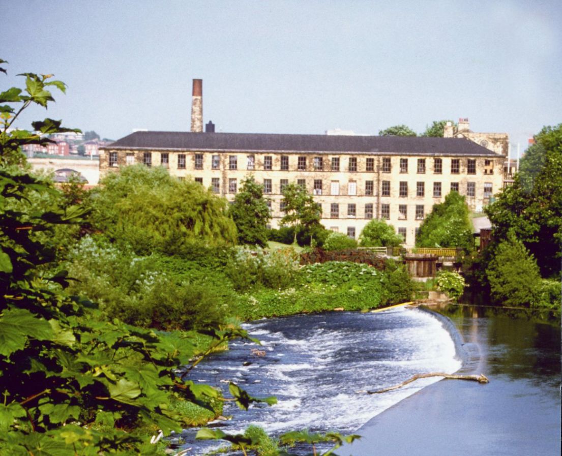 A large white building in the distance, partially hidden by trees in the foreground. There is also a sizable lake which is used by the mill for power.