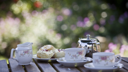 China milk jug, cups and saucers with metal tea pot and plate of scones on a table outside.