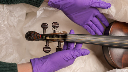 Purple latex gloved hands lift the neck of a violin away from white packaging.