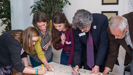 Three women and two men leaning over a table, working on a large sheet of paper.