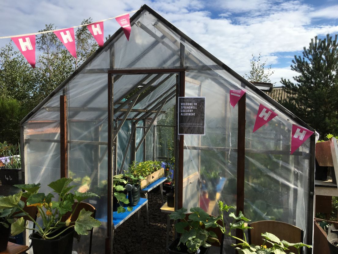 A timber structured greenhouse with seedlings potted inside, seen through the open door and see-through material. HODs bunting is hung out front.